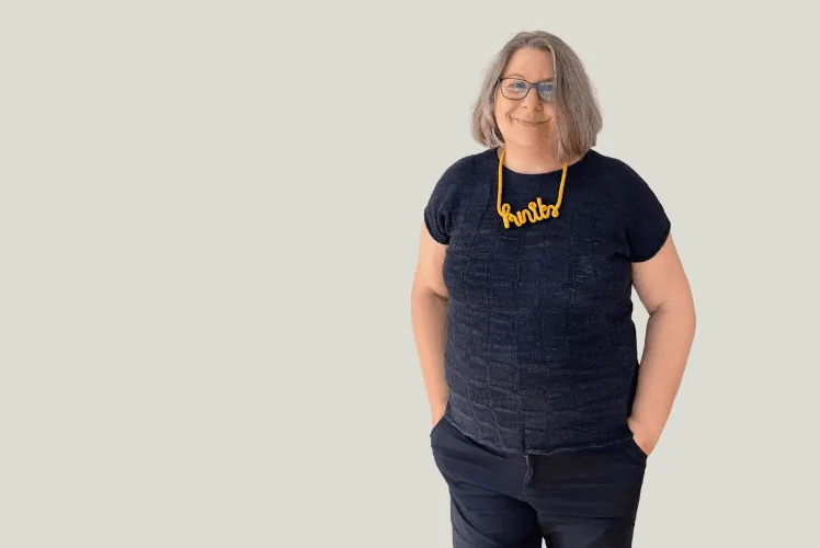 Claudia mit dunkelblauem Strickpulli mit kurzen Ärmeln, blauer Hose und gelber Kette mit Schriftzug knits