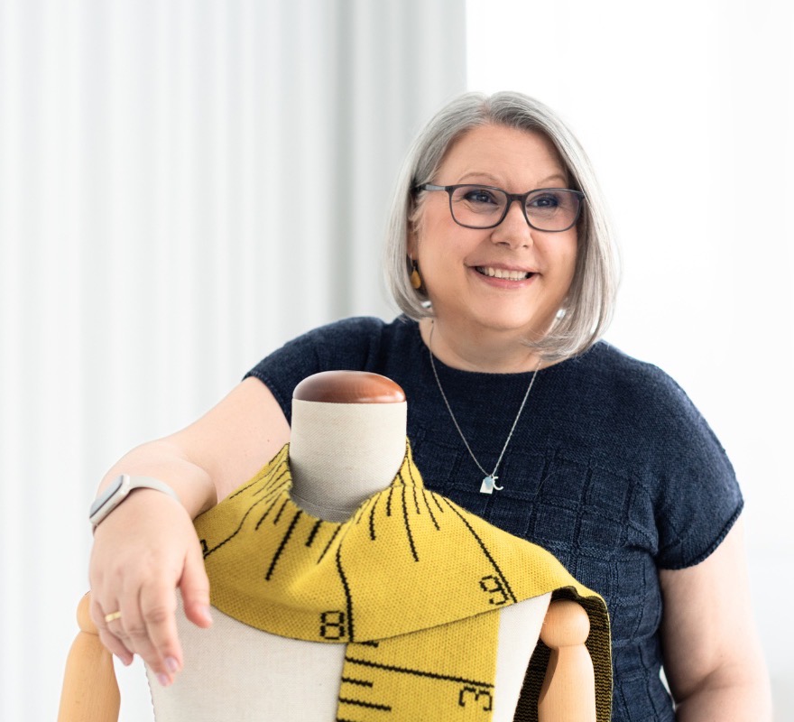 Claudia Eisenkolb lehnt lachend an einer Schneiderpuppe, über der ein gelbes Strickteil mit Maßband-Muster liegt
