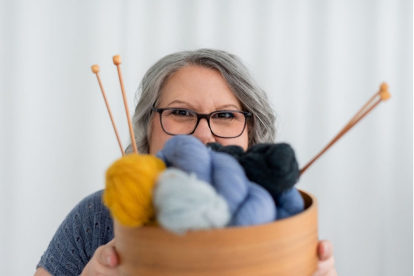 Claudia Eisenkolb hinter einer Schale mit Wollknäueln und Stricknadeln und mit einem Stapel gestrickter Pullover im Arm