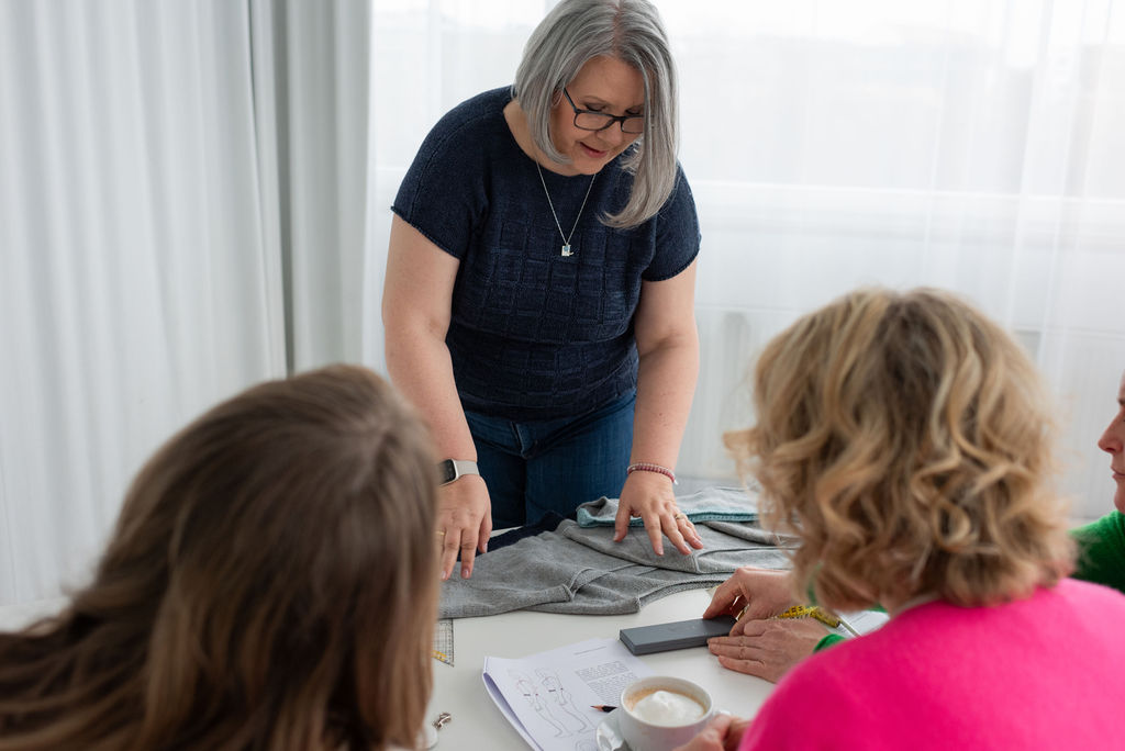Claudia steht im Workshop am Tisch und erklärt drei Teilnehmerinnen ein graues Oberteil, davor liegen Arbeitsblätter mit Schemazeichnungen