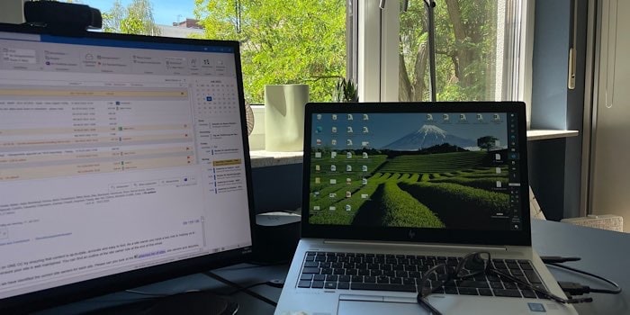 Blick aus dem Arbeitszimmerfenster mit grünem Bäumen im Hintergrund und geöffnetem Firmenlaptop rechts und zweitem großen Monitor und zweiter Tastatur links im Bild