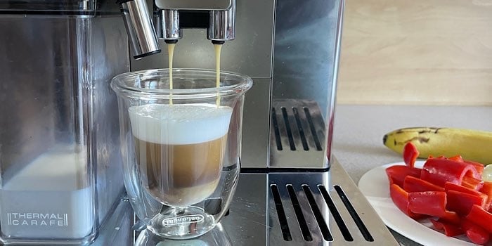 Cappuccino läuft aus einer Espressomaschine mit Milchbehälter und rechts davon die Küchenarbeitsplatte mit einem Teller mit roten Paprikastücken und einer Banane