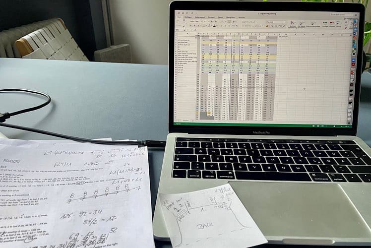 offenes MacBook Pro auf Schreibtisch mit geöffnetem Excel-Sheet zur Berechnung von Größen für ein neues Strickdesign sowie Ausdrucke der Anleitung mit handschriftlichen Notizen