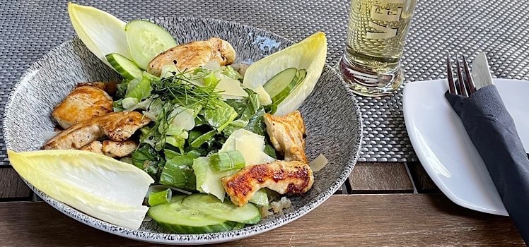 Tisch mit Teller, auf dem ein Cesar Salad mit furztrockenem Huhn angerichtet ist, daneben ein Teller mit Besteck, das in einer Serviette eingewickelt ist und ein Bierglas mit Radler
