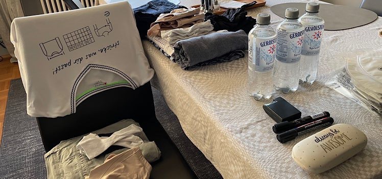 Tisch mit Tischdecke, auf der 3 Wasserflaschen stehen Wäsche, Sonnebrillenetui, zwei Stifte und ein Maßband liegen. Links daneben steht ein Stuhl über dem ein T-Shirt hängt mit dem Aufdruck: stricke, was dir passt