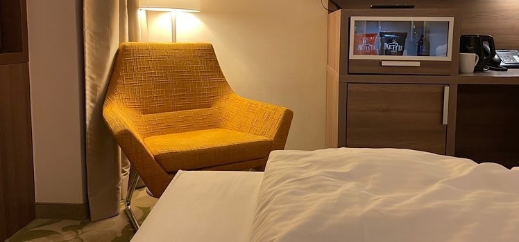 Hotelzimmer Fußende vom Bett und gelbem Sessel mit Stehlampe dahinter und Minibar daneben